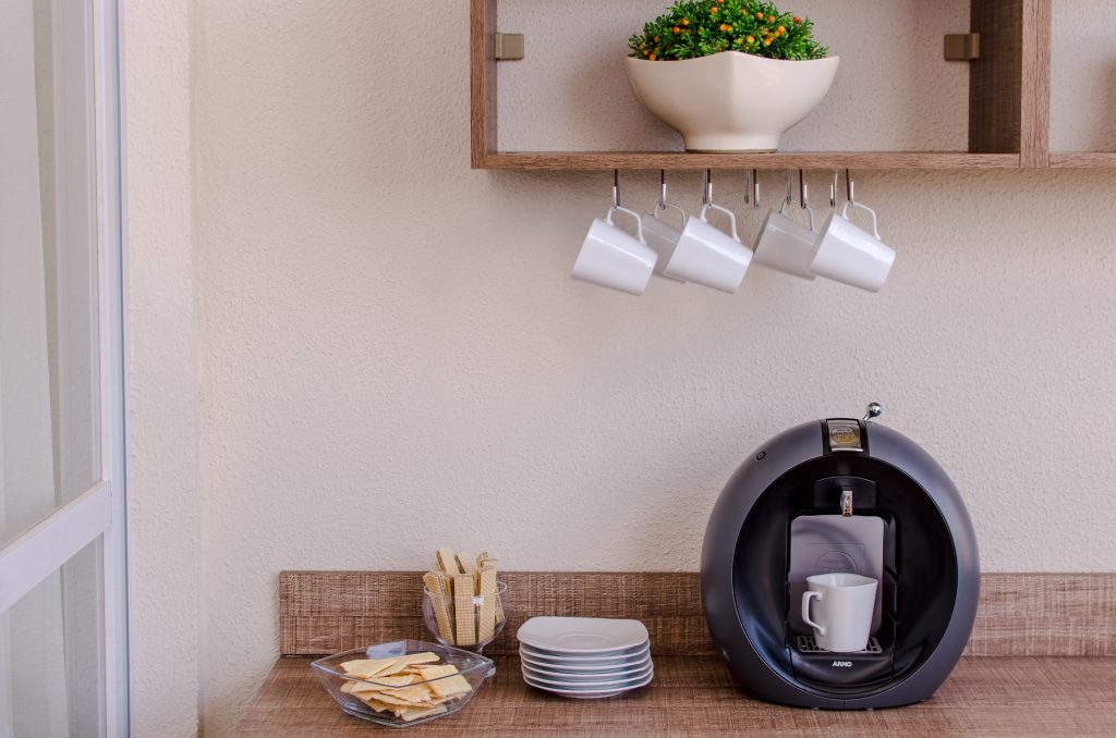 Na imagem, uma cafeteira com bolachas e pires. Ainda, em cima, xícaras em um porta-xícaras e uma planta em um prateleira.